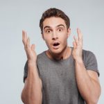 Portrait of a surprised young man holding hands at his face over white background