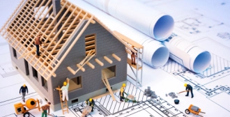 A model of a partially built house, complete with miniature workers and tools, sits atop rolled blueprints, representing the stages of home construction.