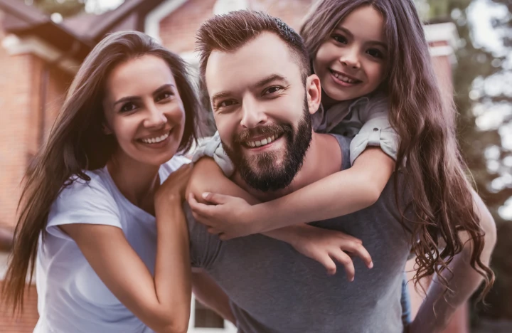 : A family of three standing in front of a house, with the father carrying his daughter on his back.