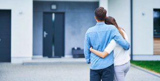 A man and woman, viewed from behind, stand arm-in-arm facing the entrance of a sleek, modern house, indicating a moment of reflection or new beginnings.
