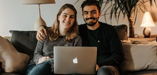 A man in a black long sleeved shirt has his arm around a woman with a grey long sleeved shirt. There is a laptop in front of them.