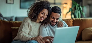  A woman in a sweater has her arm around a man. They are both looking at a laptop.