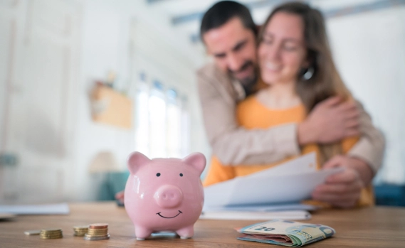A couple embracing in the background with a focus on a piggy bank, coins, and cash on a table, symbolising savings or financial planning.