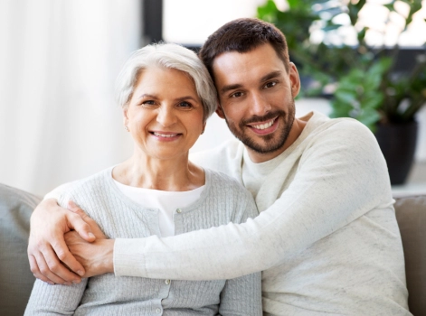 Young man embracing his elderly mother on a sofa, both smiling at the camera, conveying family warmth and closeness.