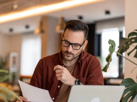 A man looking at a paper while having his laptop open in front of him