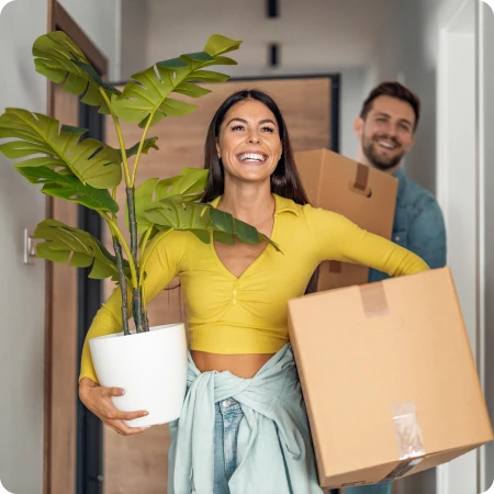 Smiling couple carrying large carton boxes and a potted plant into a new home.