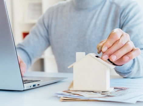A person in a grey sweater is placing a coin into a small wooden house model while using a laptop. The scene suggests financial planning or investment, with papers and documents spread out on the desk.