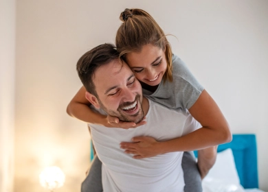 A cheerful couple shares a joyful moment, with the woman playfully riding on the man's back. They are both smiling and laughing, creating a warm and happy atmosphere in what appears to be their home.