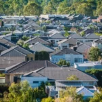An aerial view of a suburban neighbourhood featuring a variety of modern houses with tiled roofs and lush greenery. The dense cluster of homes is surrounded by trees, creating a picturesque and serene residential area.
