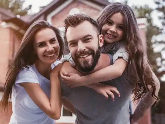 : A family of three standing in front of a house, with the father carrying his daughter on his back.