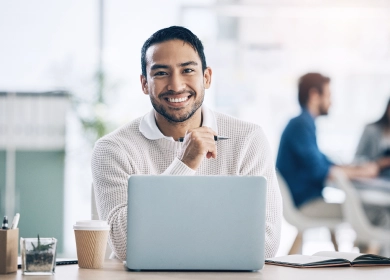 A smiling man wearing a white sweater sits at a desk with a laptop, coffee cup, and notebook. He appears to be in a modern office setting, conveying a sense of productivity and enthusiasm in a collaborative work environment.