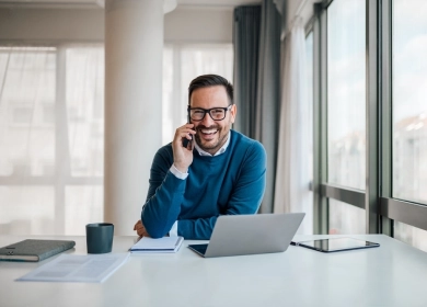 A cheerful man wearing glasses and a blue sweater is sitting at a desk, talking on his mobile phone. He has a laptop, notebook, and tablet in front of him, suggesting a productive work environment in a bright, modern office space.