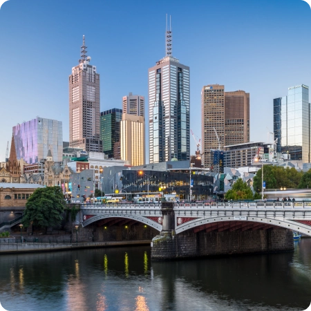 Melbourne city skyline at dusk featuring iconic landmarks, including Flinders Street Station, St. Paul's Cathedral, and modern skyscrapers, with reflections shimmering in the calm waters of the Yarra River.
