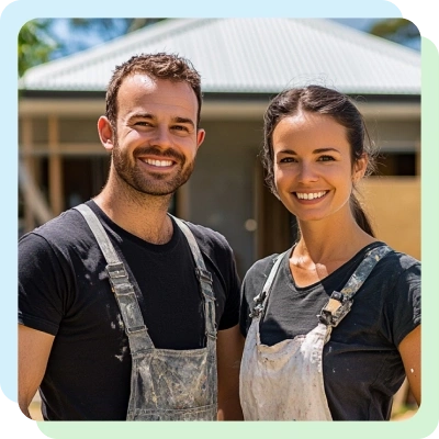 A smiling couple in paint-splattered overalls stands in front of a partially constructed house