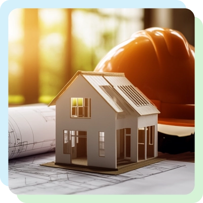 A miniature model of a house sits on architectural blueprints, with a bright orange construction helmet in the background