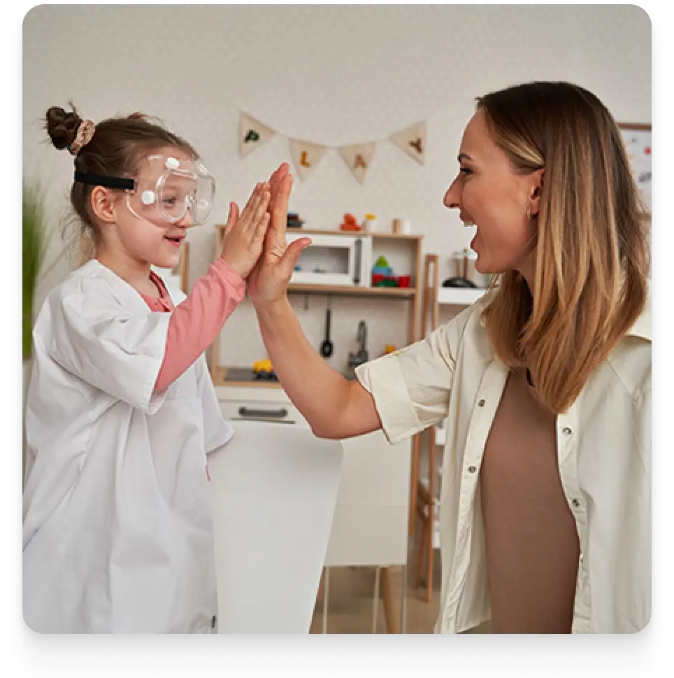 A woman in a white coat and a girl are smiling and giving each other a high five indoors, against a wall. Both appear joyful and are dressed casually.
