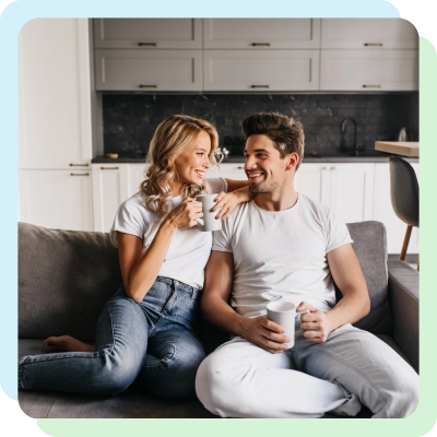 A couple sitting together on a couch, smiling and enjoying coffee in a modern kitchen setting.