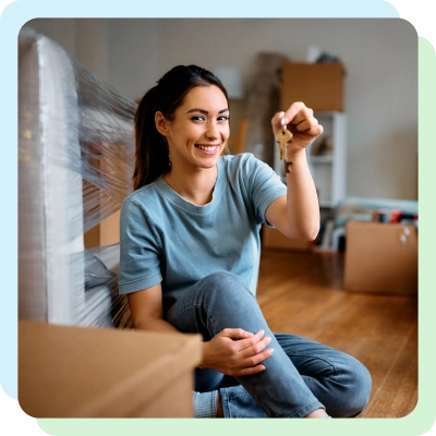 A girl sits on a wooden floor amidst moving boxes, holding up a set of keys with a smile, indicating a new home.