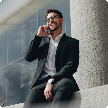 A man in a suit and glasses is talking on a cell phone while standing outdoors against a wall.