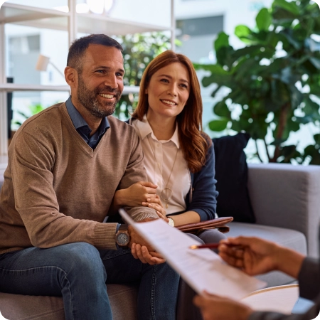 A couple sits closely on a sofa, holding hands, while a person across from them discusses paperwork in a modern office setting.
