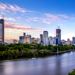 Brisbane cityscape at dusk, featuring tall skyscrapers under a colorful sky, with a tree-lined riverbank and sailboats in the foreground.