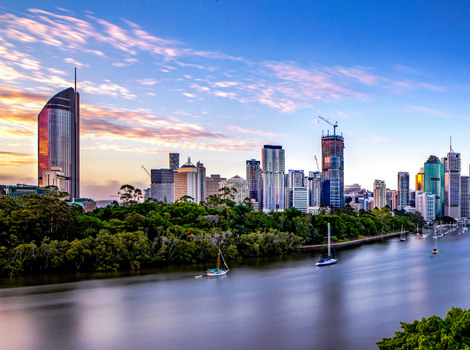 Brisbane cityscape at dusk, featuring tall skyscrapers under a colorful sky, with a tree-lined riverbank and sailboats in the foreground.