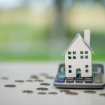A small model home sits on top of a calculator, with coins scattered around it.
