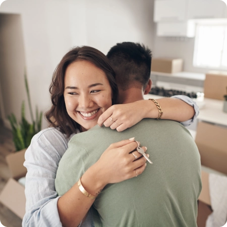 A smiling couple stands in a room with moving boxes; the woman holds a key, suggesting they have just moved into a new home.