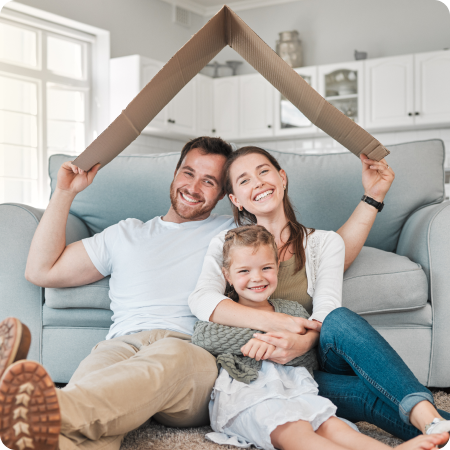 First home buyers sitting together on a sofa, smiling while holding a cardboard house shape above their heads