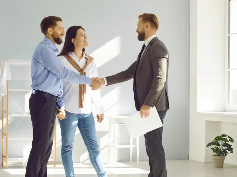 Couple greeting a suited man with a handshake in a sunlit room.